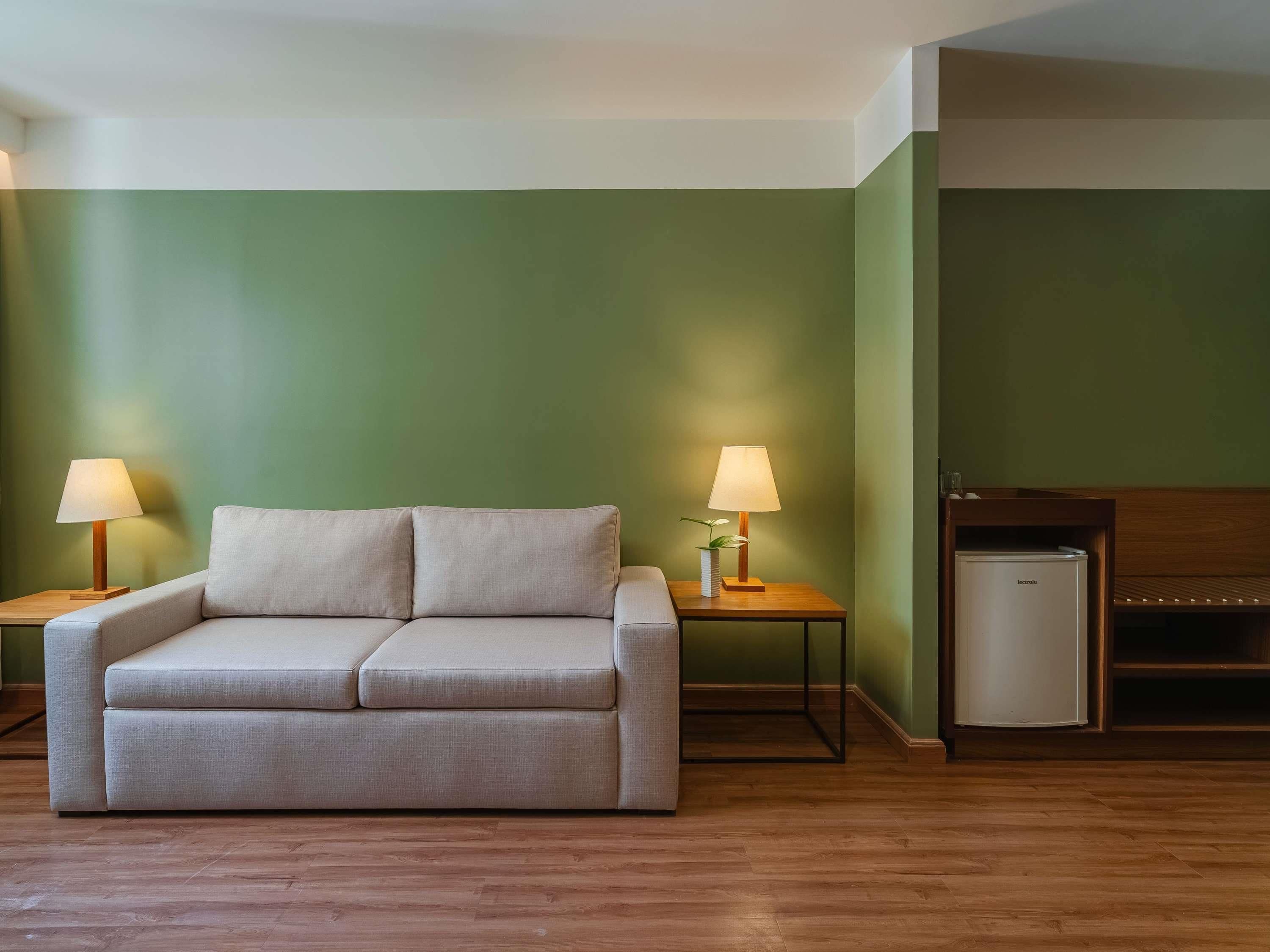 A room with a sofa, two bedside tables with lamps, a small refrigerator, and a wooden bench.