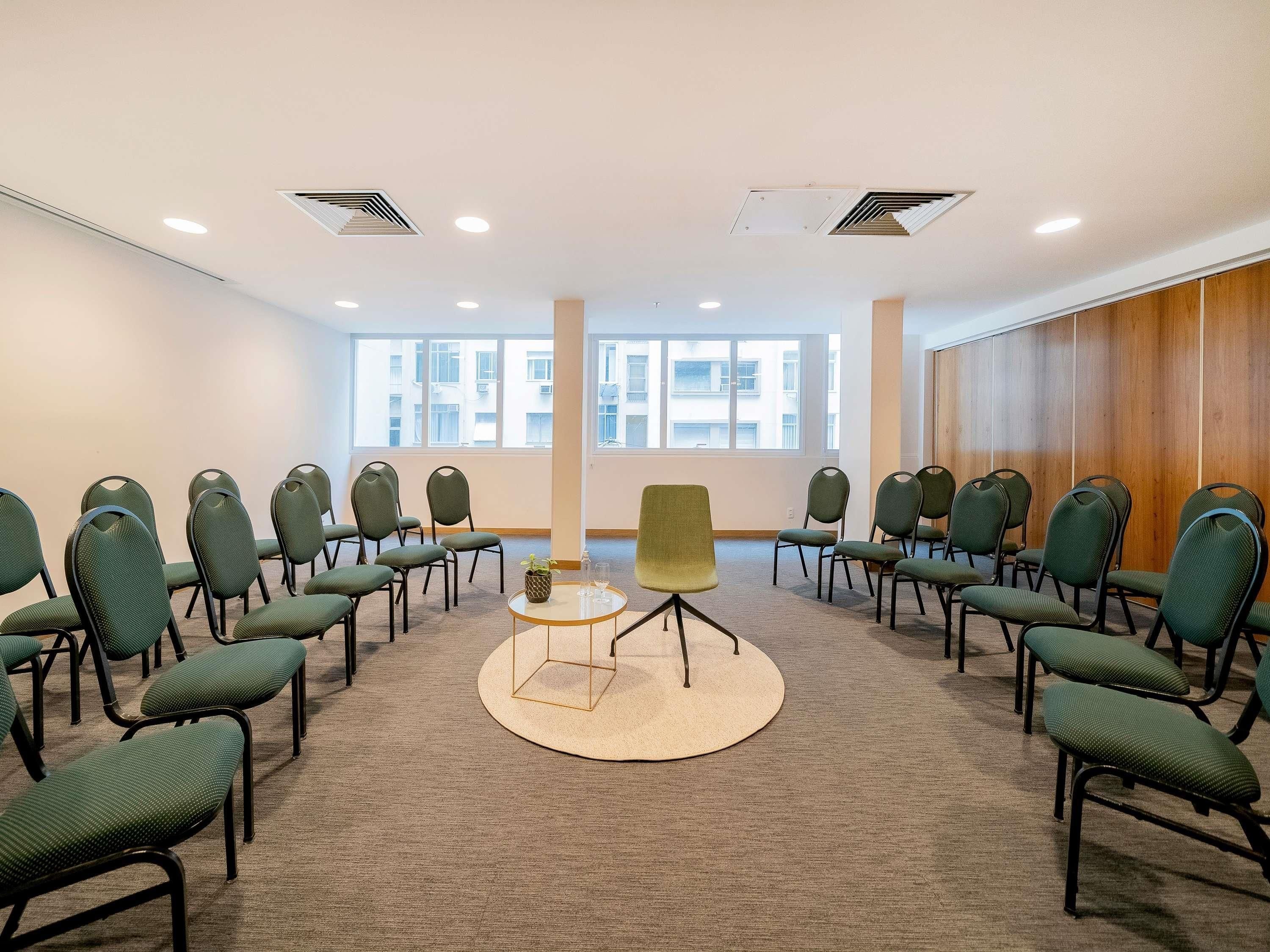 Une salle de conférence avec des rangées de chaises vertes, une table centrale et de grandes fenêtres.