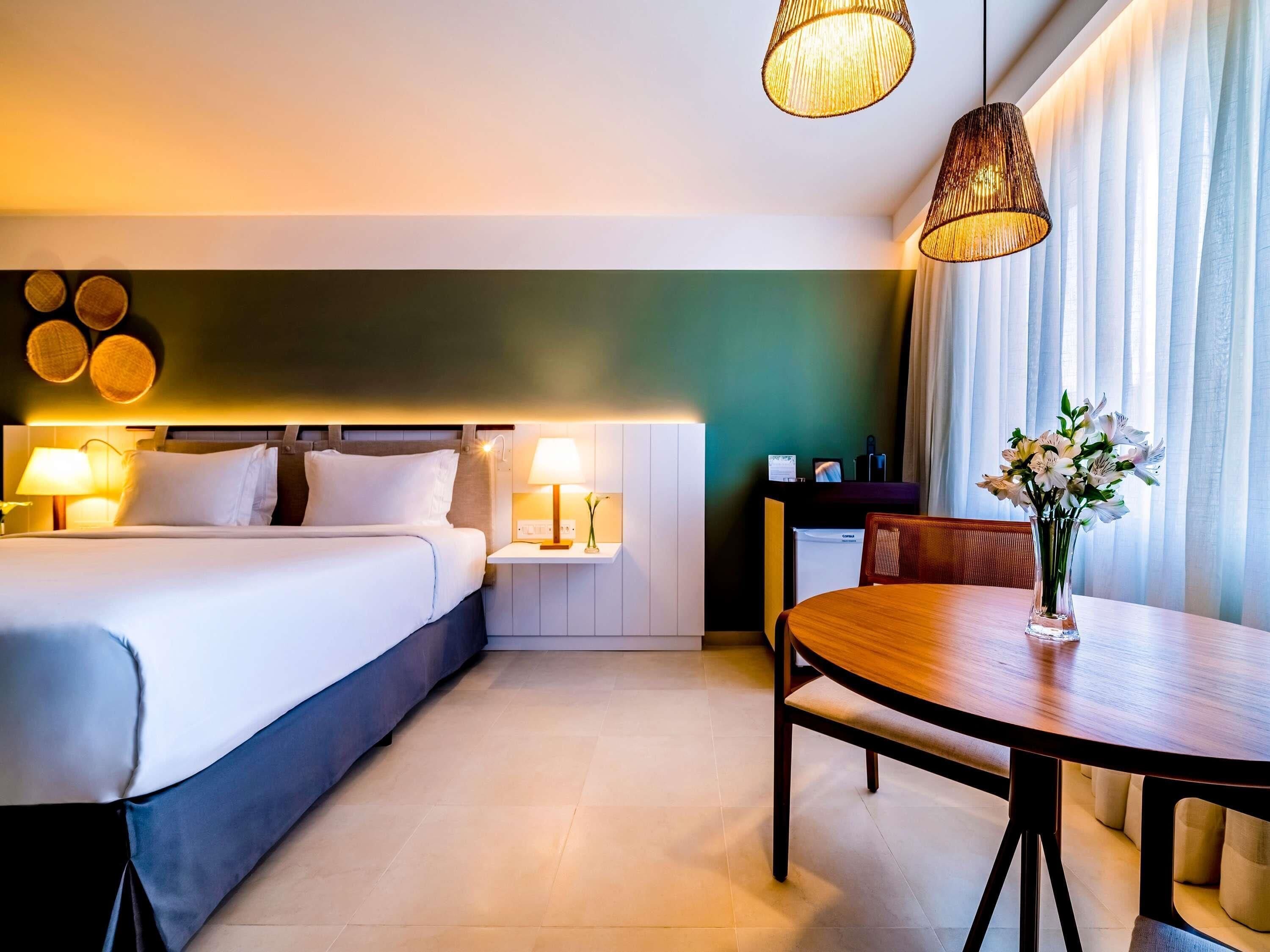 Une chambre d’hôtel comprenant un lit, des tables de chevet avec des lampes, une table à manger avec des chaises et un vase avec des fleurs.