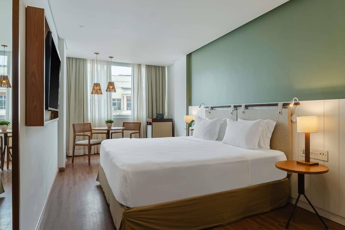A hotel room with a bed, a desk, a chair, a TV, and a lamp.