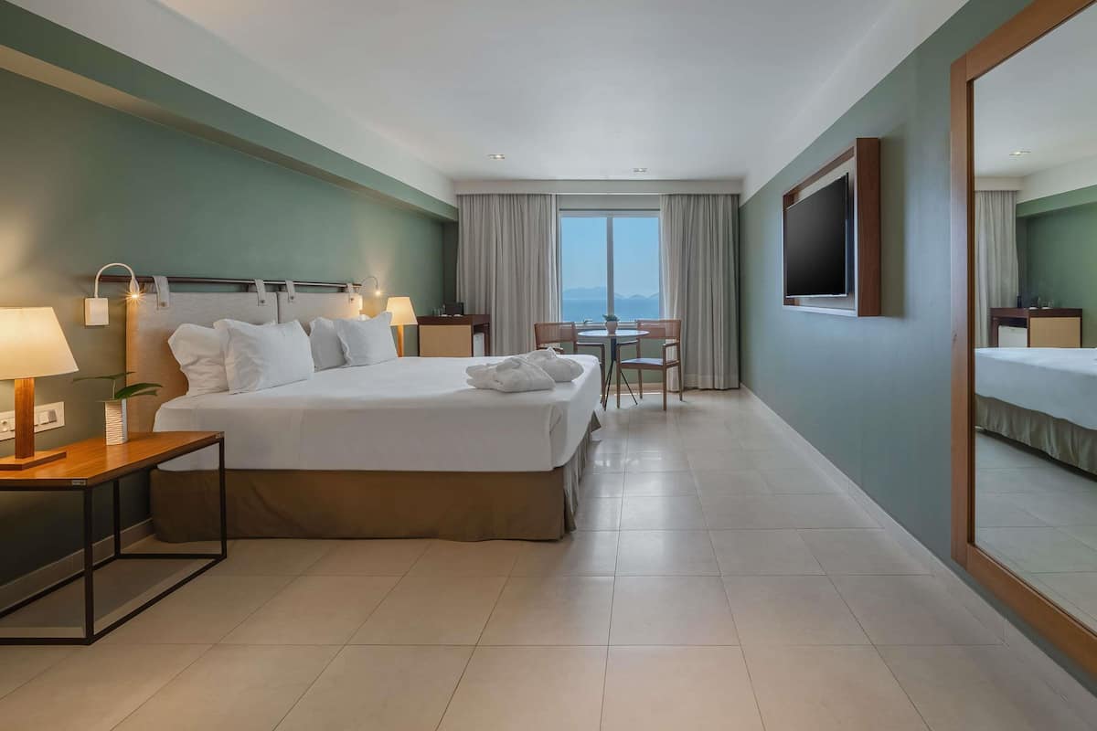 A hotel room with a bed, bedside tables, a desk, a chair, a TV, and a large window.