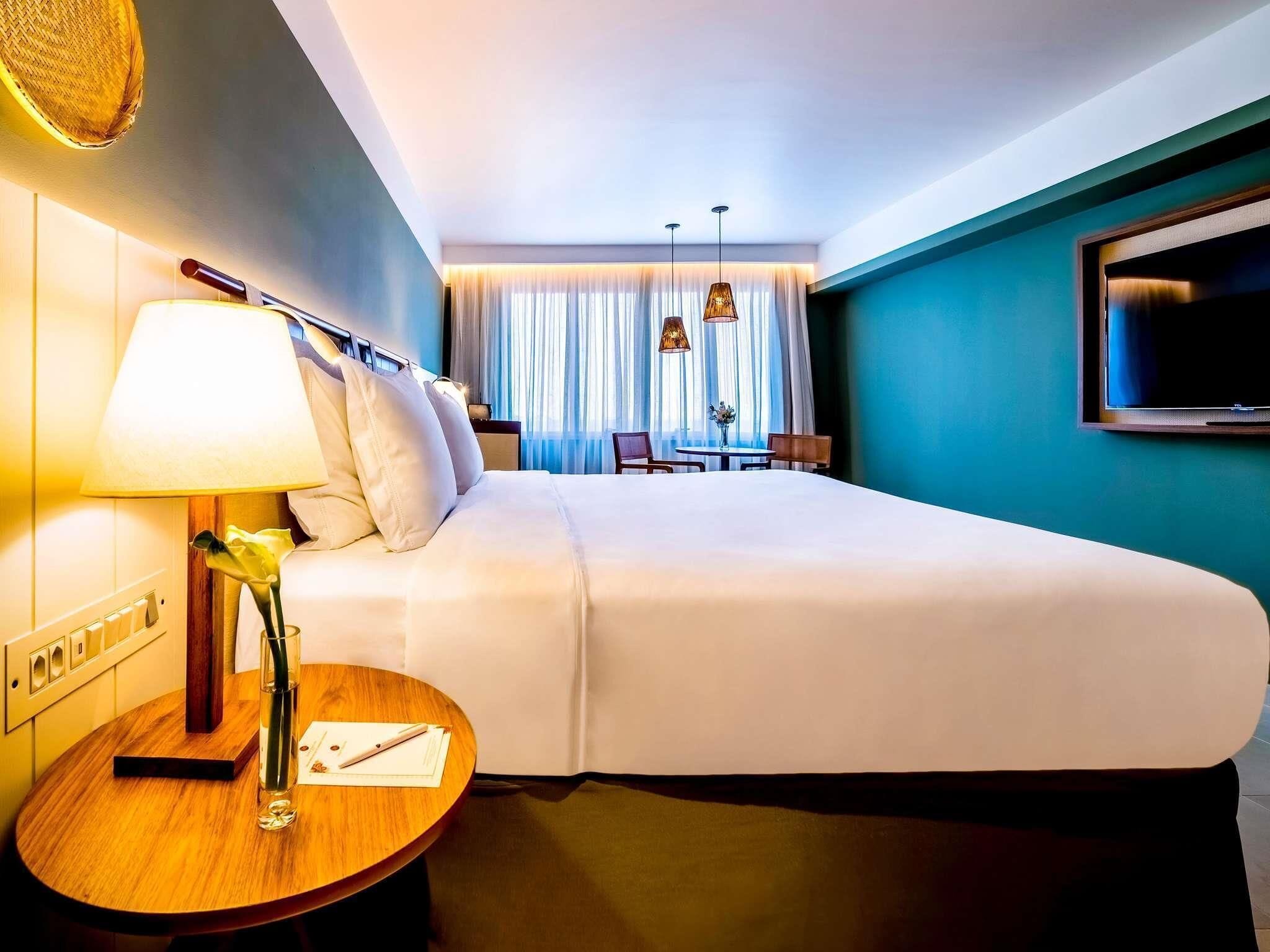 Quarto de hotel com uma cama grande, uma mesa de cabeceira com abajur, televisão na parede e vista do interior do quarto.
