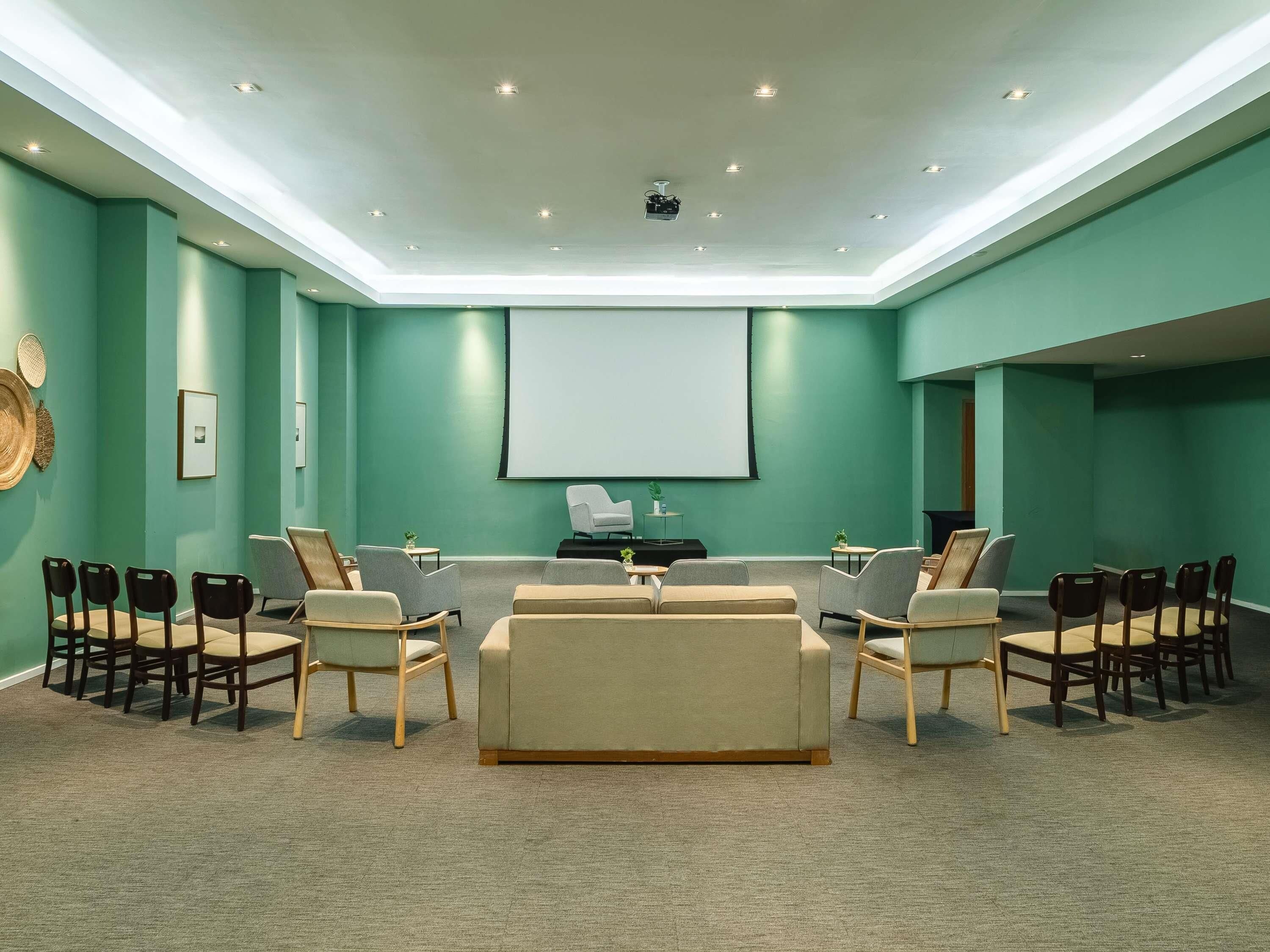 Une salle de conférence aux murs de couleur turquoise, dotée d’un grand écran et de nombreuses chaises disposées autour d’une table centrale.