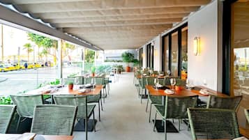 An outdoor dining area with wicker chairs and tables, large windows, and potted plants.