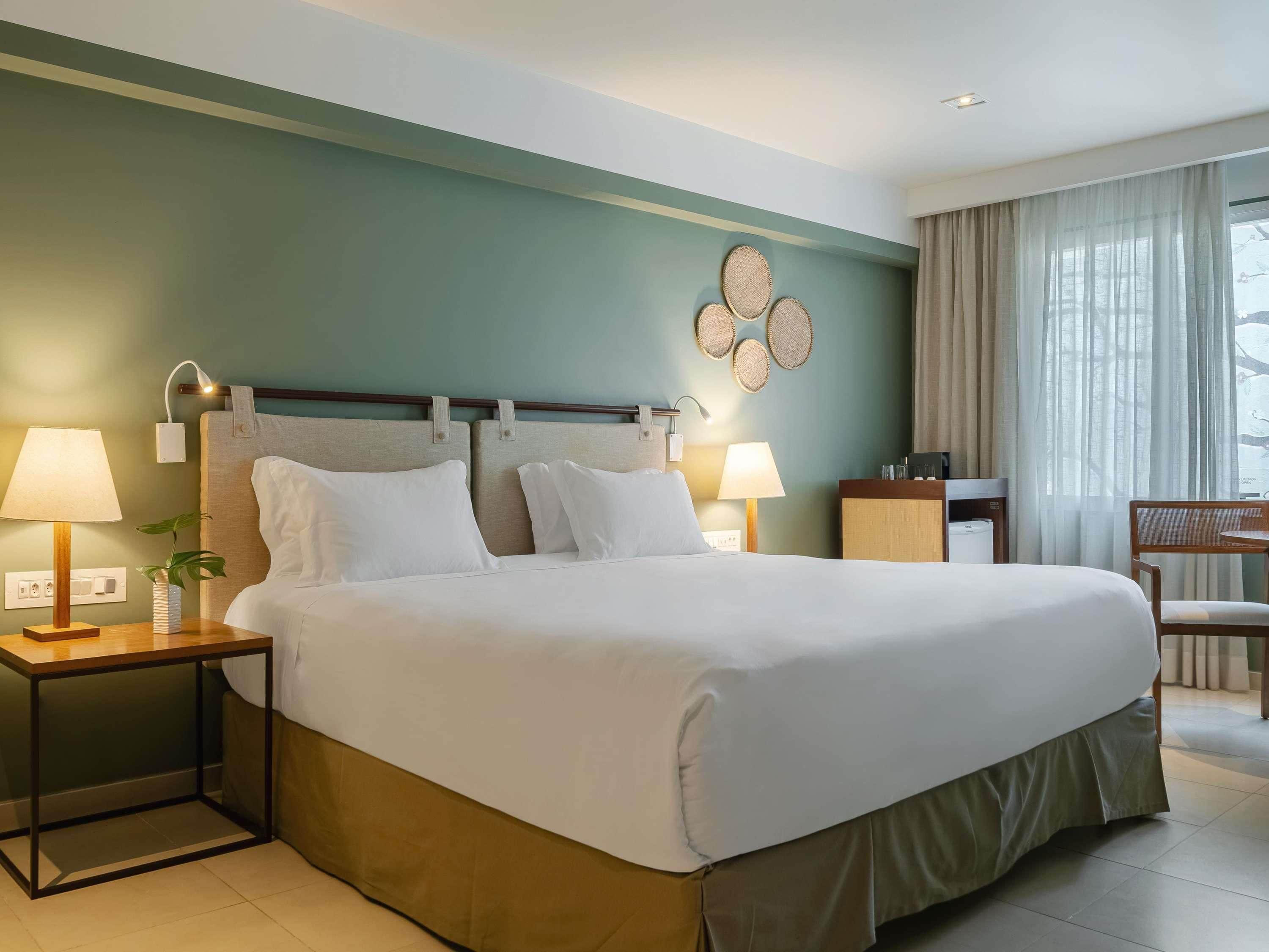 Hotelkamer met een groot bed, twee nachtkastjes met lampen, een bureau met stoel, en uitzicht op het omliggende landschap door dunne gordijnen.