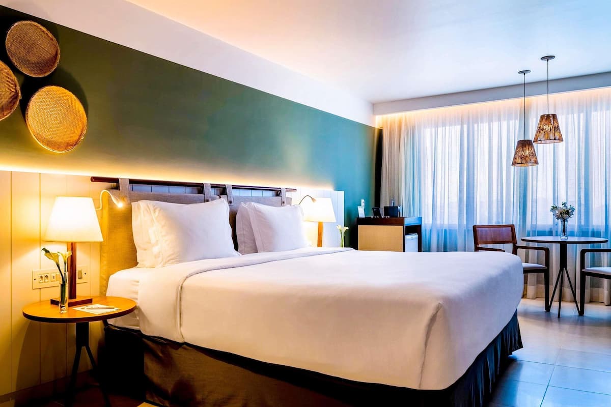 A hotel room with a bed, bedside table, lamp, chair, and a small table with a vase.