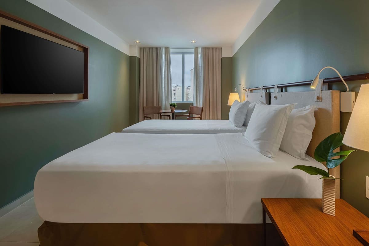 A hotel room with two beds, a TV, a desk, a chair, a lamp, and a window with curtains.