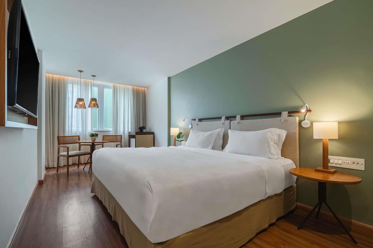 A hotel room with a large bed, a desk, chairs, a TV, and a lamp.