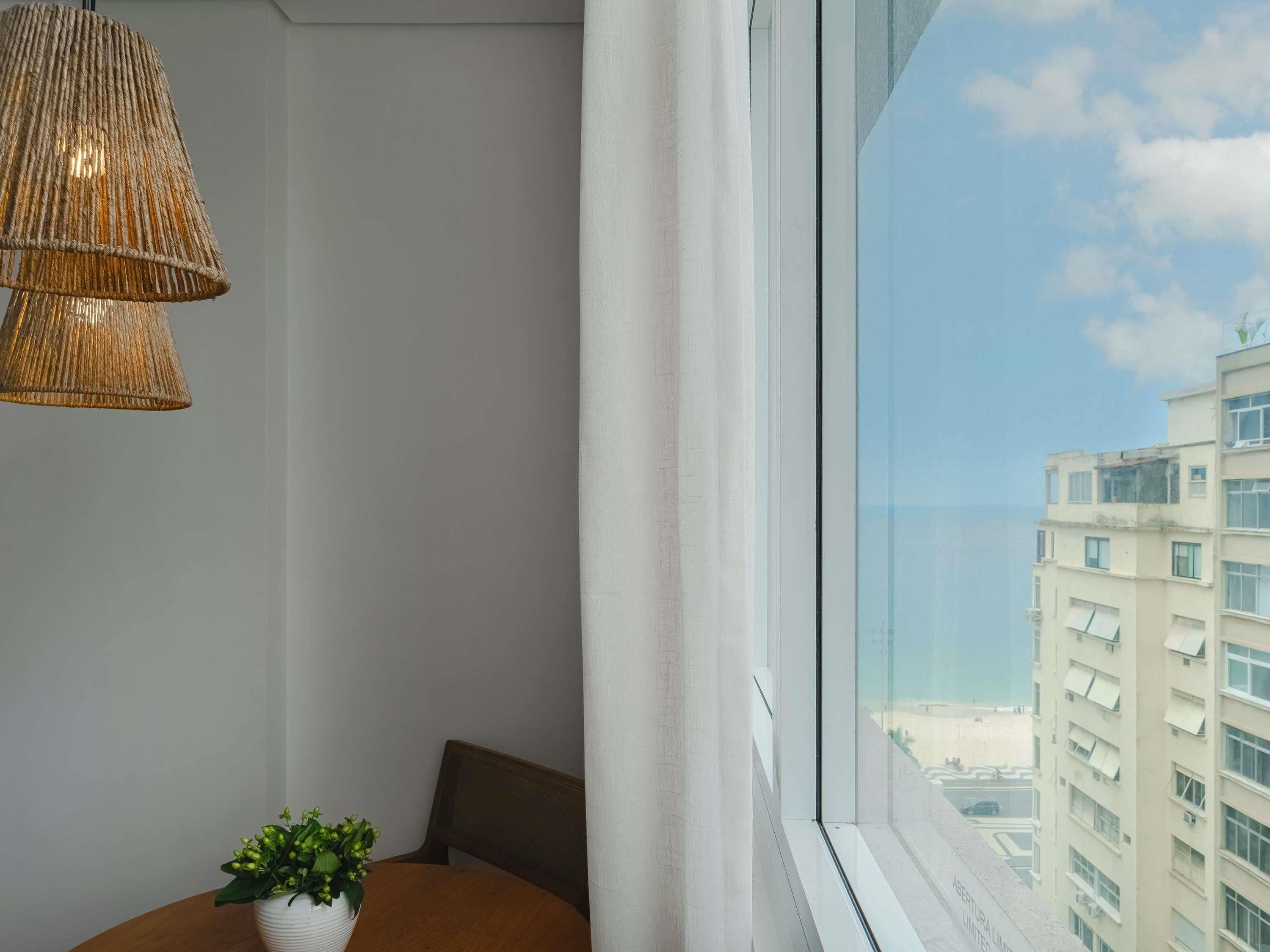 Una habitación con un ventanal grande, una lámpara de mimbre, una planta en maceta y una silla de madera.