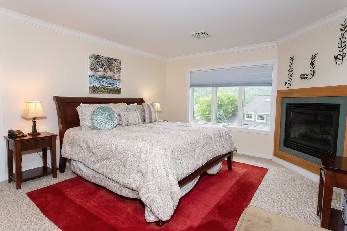 A bedroom with a large bed, two bedside tables, a red rug, a fireplace, and a window with a view of trees.