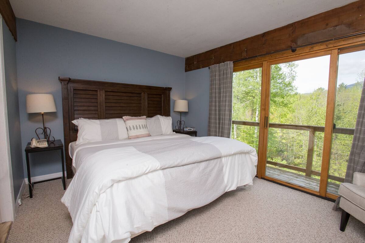 A bedroom with a large bed, two bedside tables, a nightstand, and a window with a view of greenery.