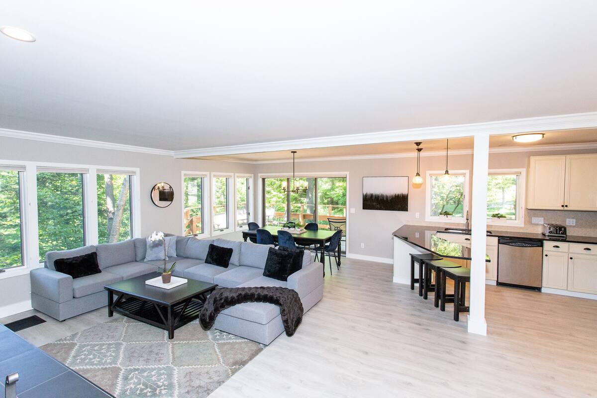 A spacious living area with a gray sofa, a coffee table, and a dining table with chairs. There is a kitchen area with cabinets and a dishwasher.