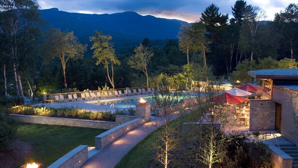 Zona de piscina con sillones de descanso y un hogar, rodeada por un jardín ajardinado y un edificio de fachada de piedra.