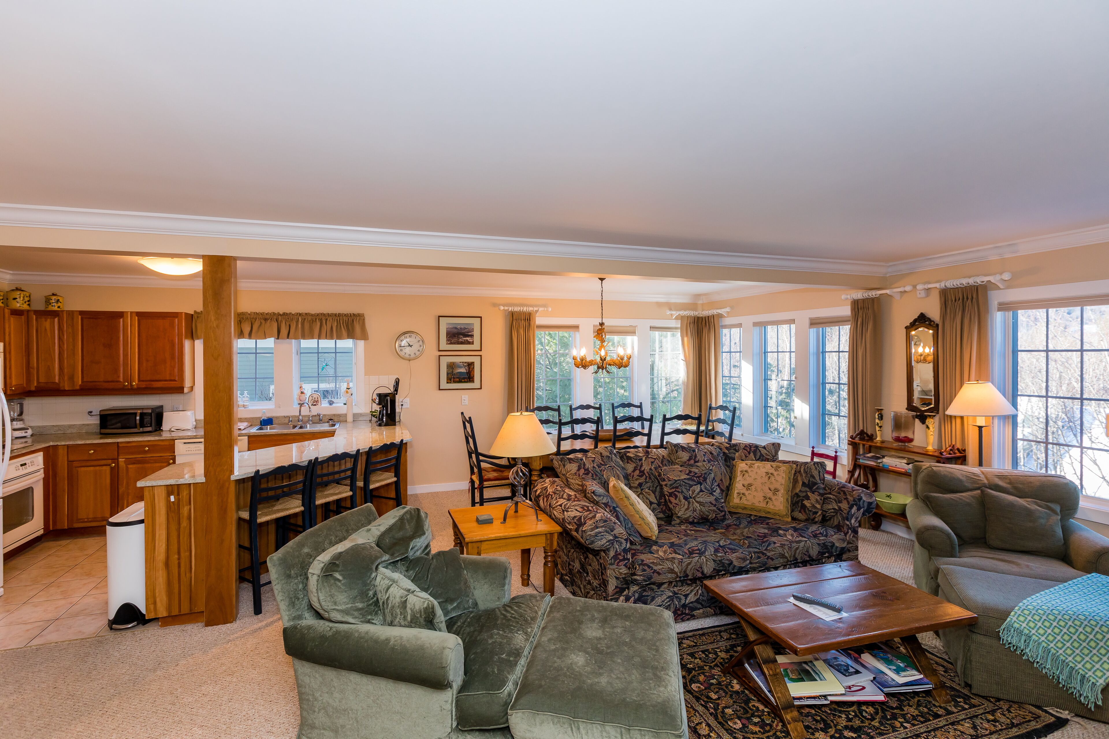 A spacious living room with a sofa, armchairs, and a coffee table. A dining area with a table and chairs is adjacent to the living room. The kitchen is visible in the background with wooden cabinets and a microwave.