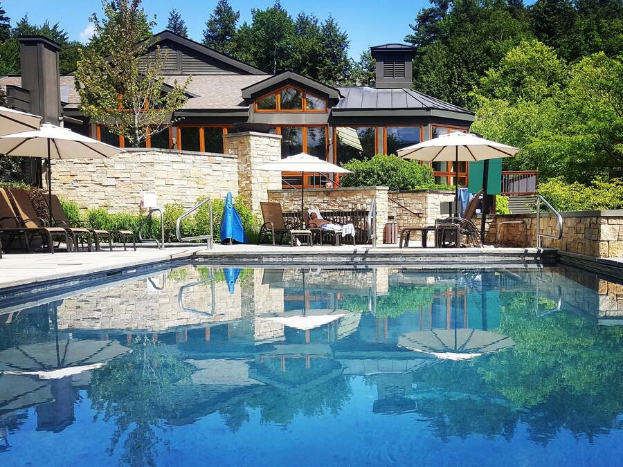 A swimming pool with lounge chairs and umbrellas, surrounded by stone walls and a large house with multiple windows.