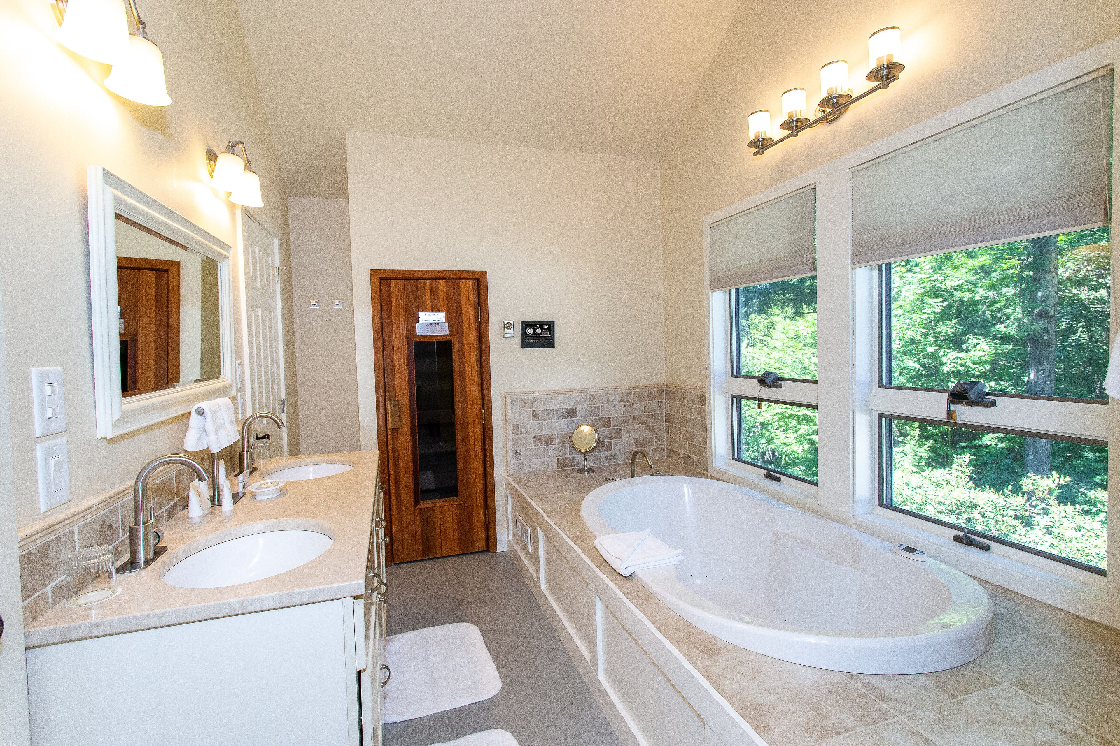 A modern bathroom with two sinks, a large bathtub, and a view of the outdoors through large windows.