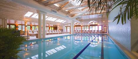 An indoor swimming pool with clear water, wooden ceiling beams, and lounge chairs along the poolside.