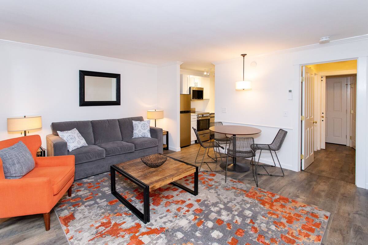 A modern living room with a grey sofa, orange armchair, and a coffee table. There is a dining area with a round table and chairs. The kitchen is visible in the background.