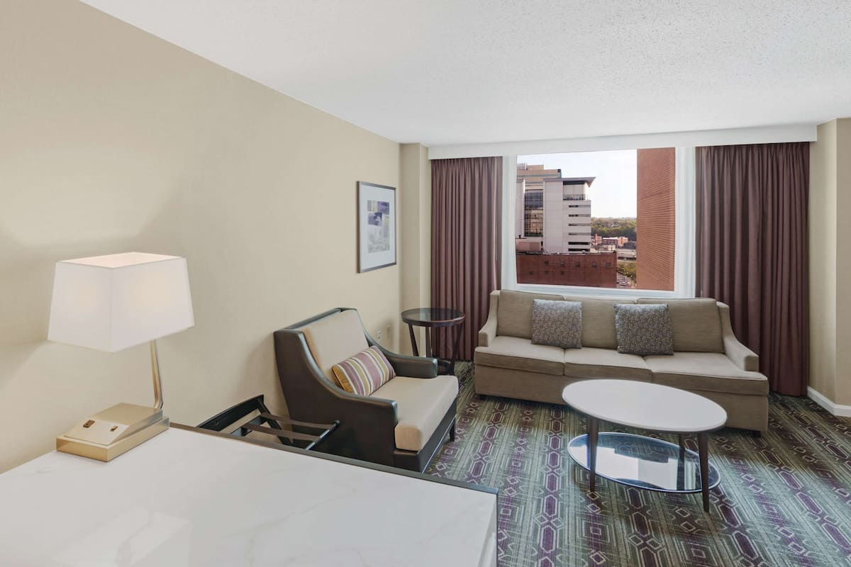A hotel room with a sofa, a small table, a lamp, and a view of the city through large windows.