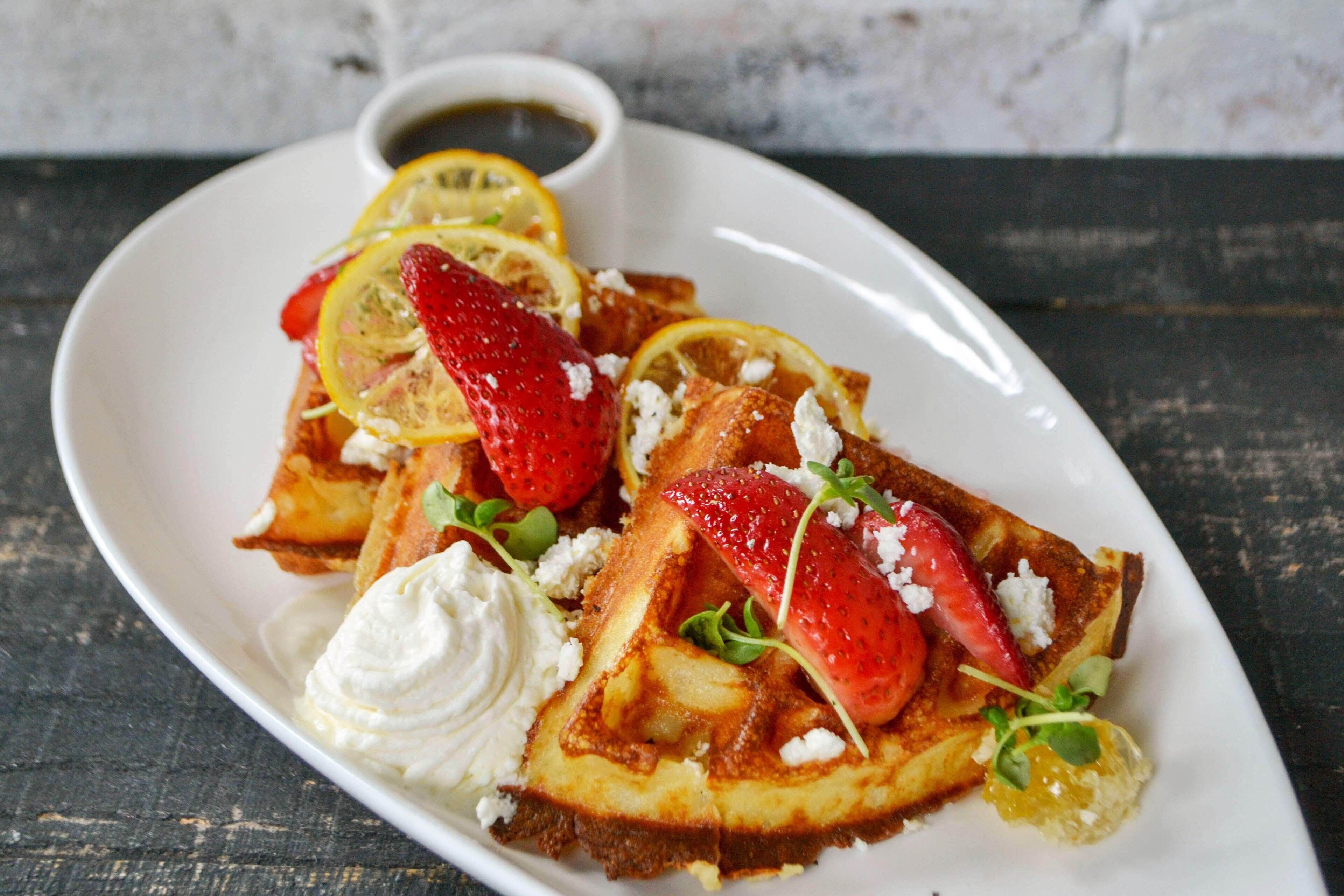 A plate of waffles topped with strawberries, lemon slices, and a dollop of whipped cream, accompanied by a small bowl of syrup.