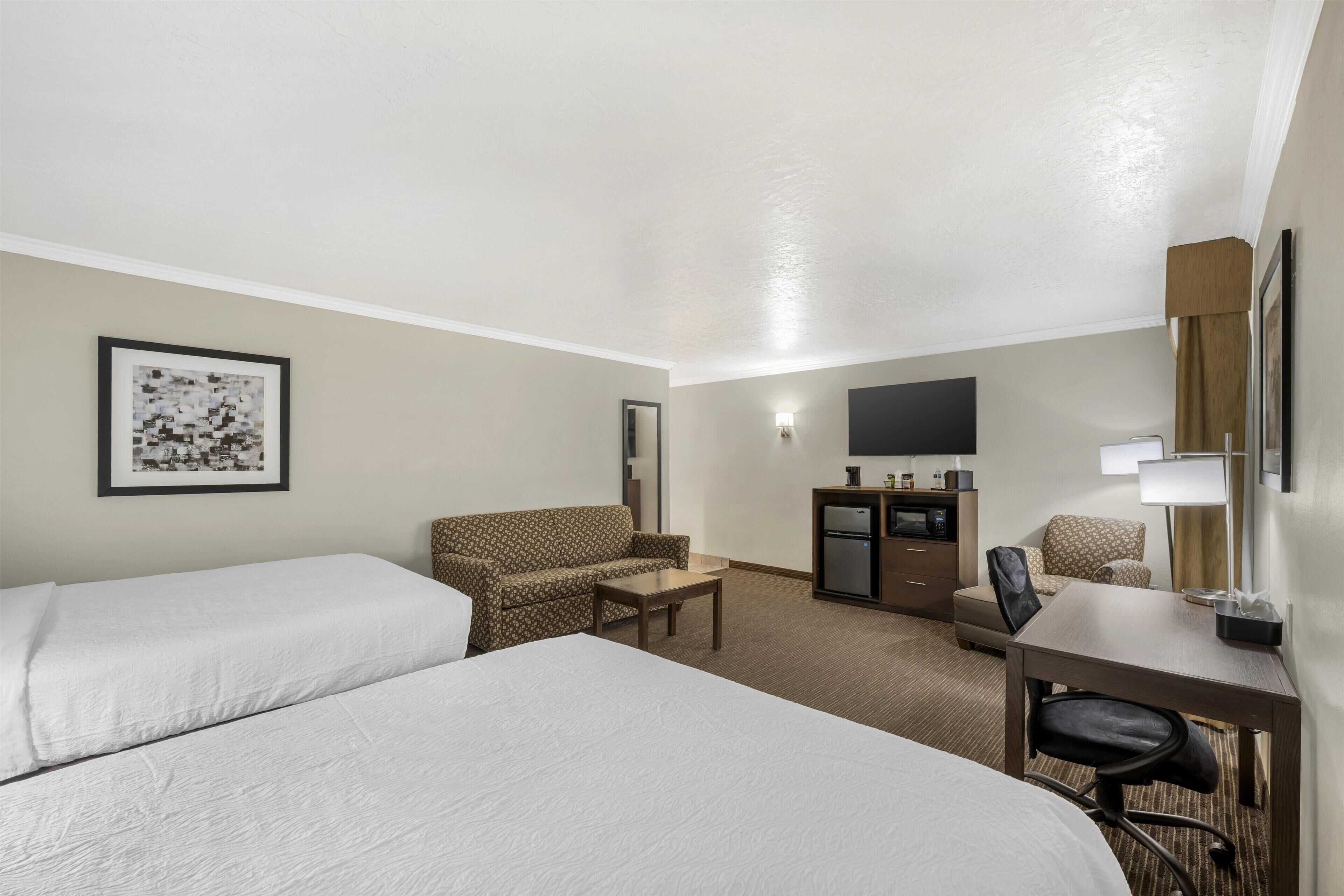 A hotel room with two beds, a desk, a chair, a sofa, a TV, and a minibar.