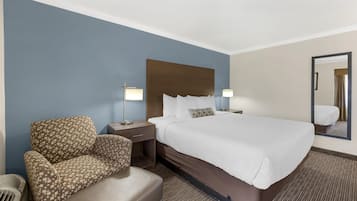 A hotel room with a large bed, a patterned armchair, a nightstand with a lamp, and a mirror on the wall.