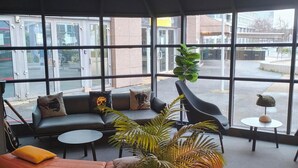 A modern lounge area with a sectional sofa, a round coffee table, and a potted plant. Large windows provide a view of the street outside. - ibis Nanterre la Défense (Nanterre)