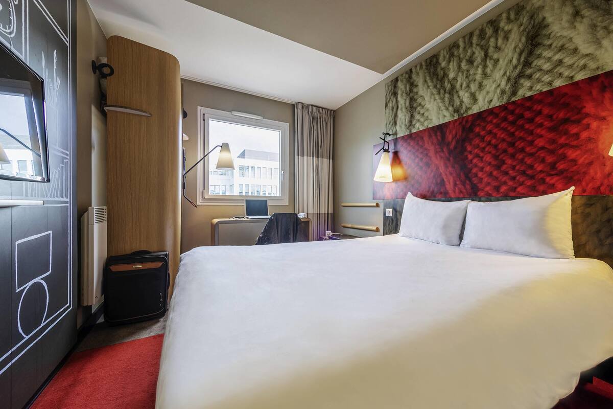 A hotel room with a large bed, a desk with a laptop, a window with a view of buildings, and a red carpet.
