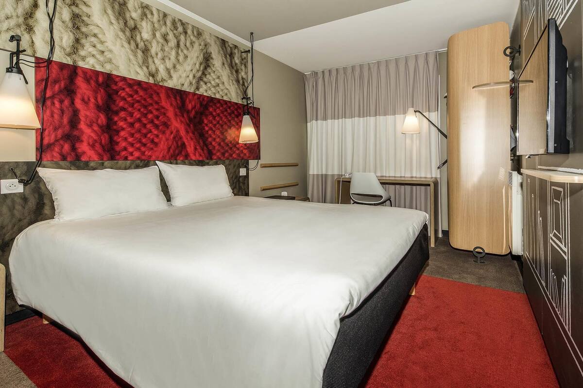 A hotel room with a large bed, a desk, a chair, a TV, and a wardrobe.