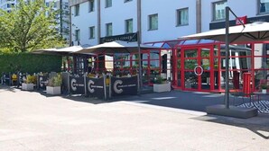 An outdoor seating area with umbrellas and tables, a red glass structure, and a sign for "Charlie’s Corner". - ibis Nanterre la Défense (Nanterre)