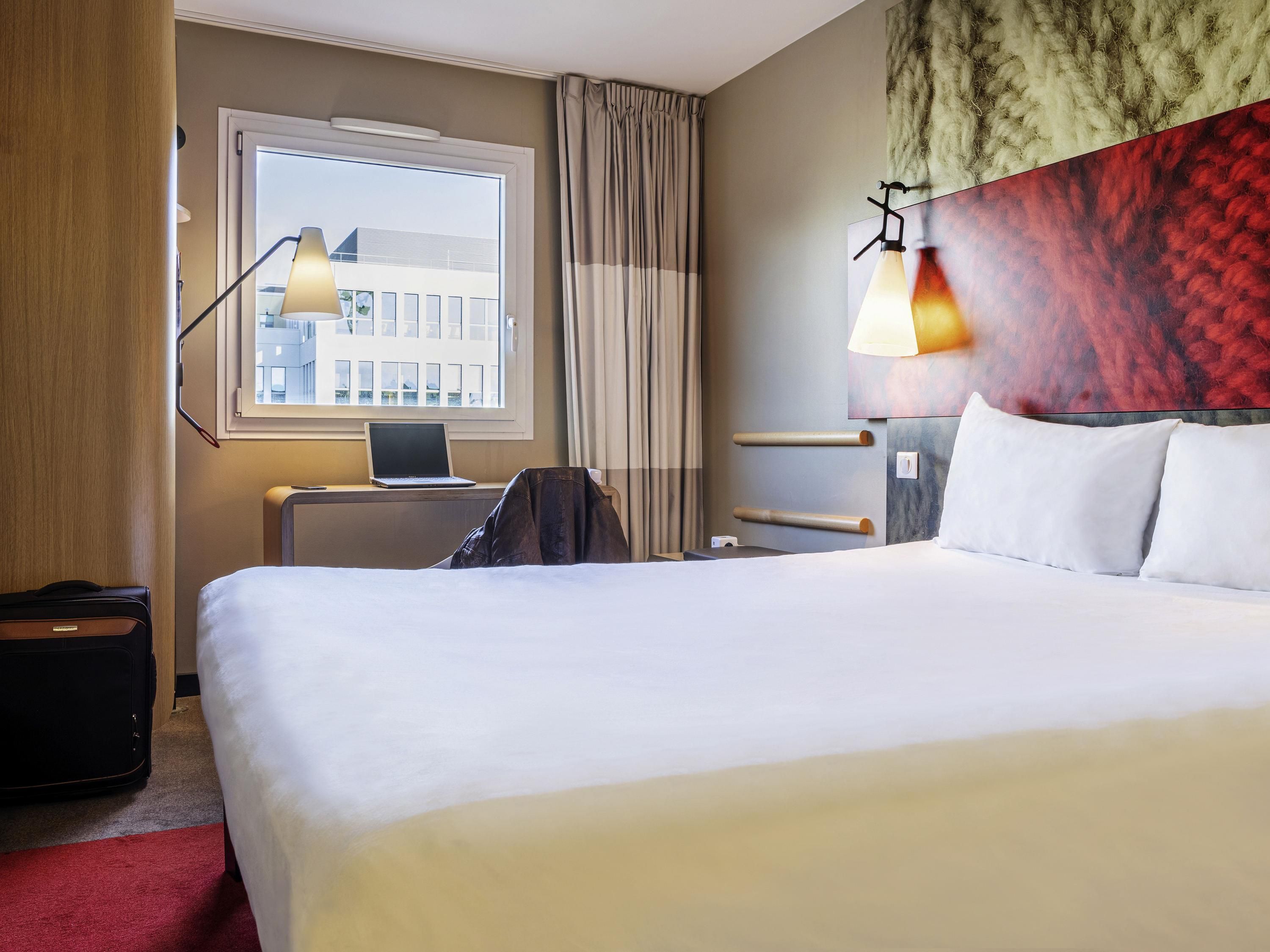 Ein Hotelzimmer mit einem Bett, einem Schreibtisch mit einem Laptop, einem Koffer, einer Lampe und einem Fenster mit Vorhängen.