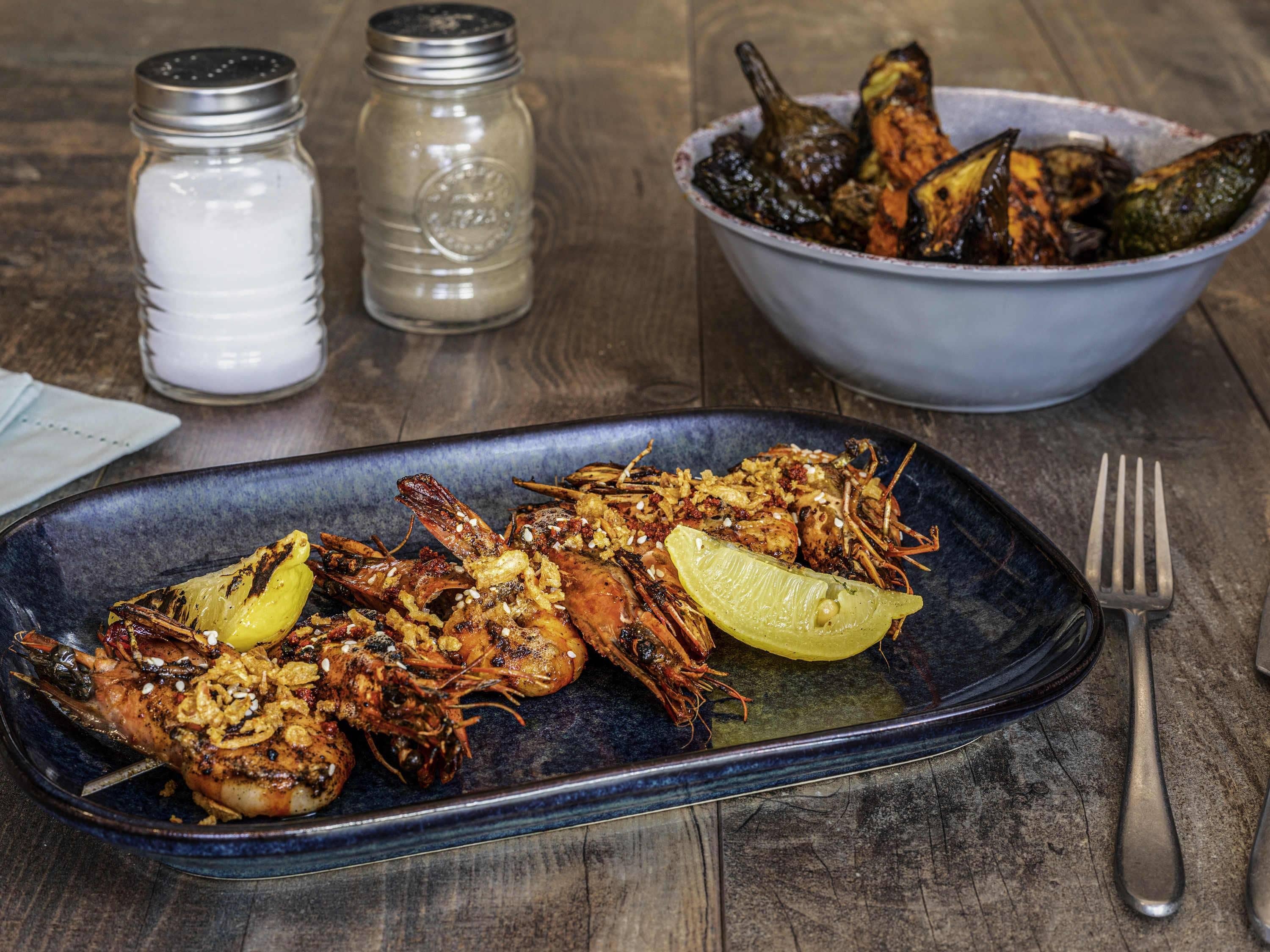 A seafood dish on a plate, a bowl of vegetables, salt and pepper shakers, and a fork on a wooden table.