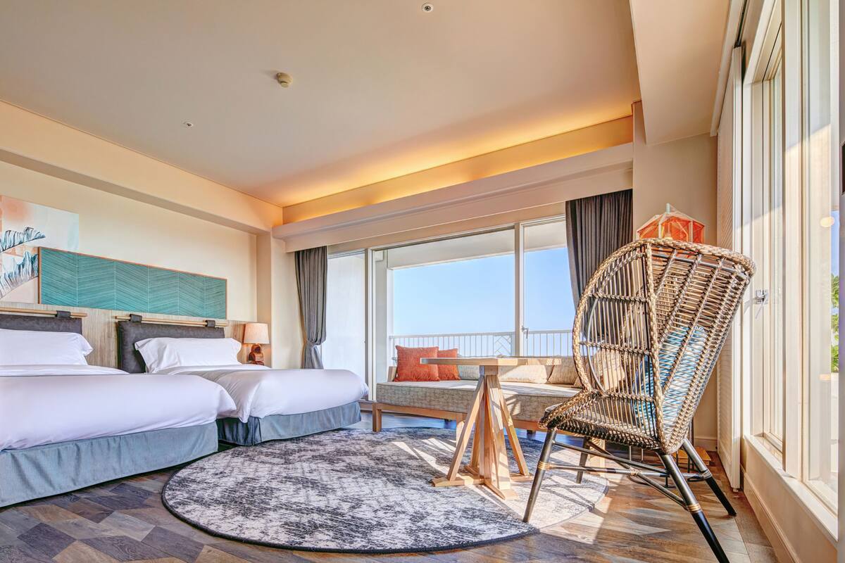 Une chambre d’hôtel avec deux lits, un fauteuil en osier, un canapé, une grande fenêtre et une vue sur l’extérieur.