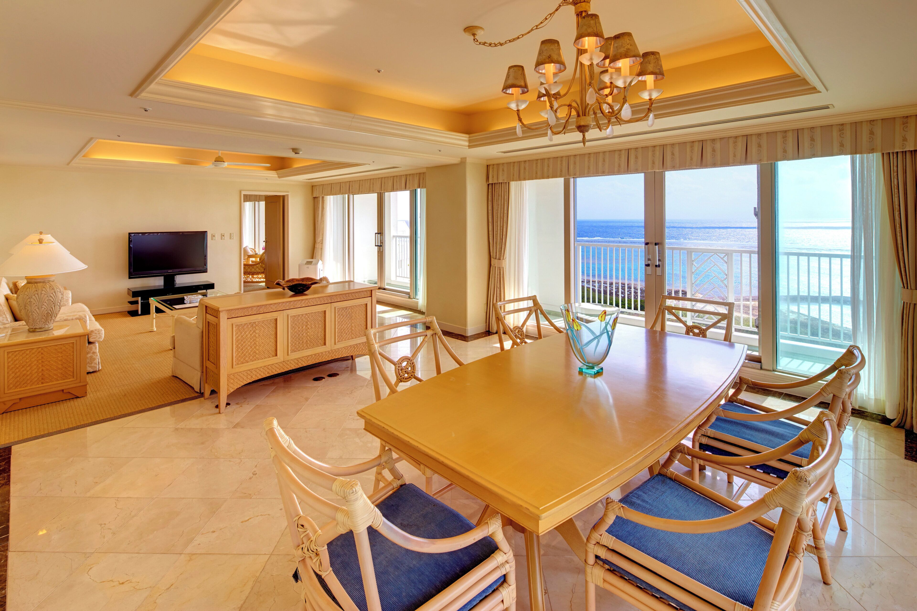 A spacious living room with a large dining table, comfortable seating, a television, and a view of the ocean through large windows.