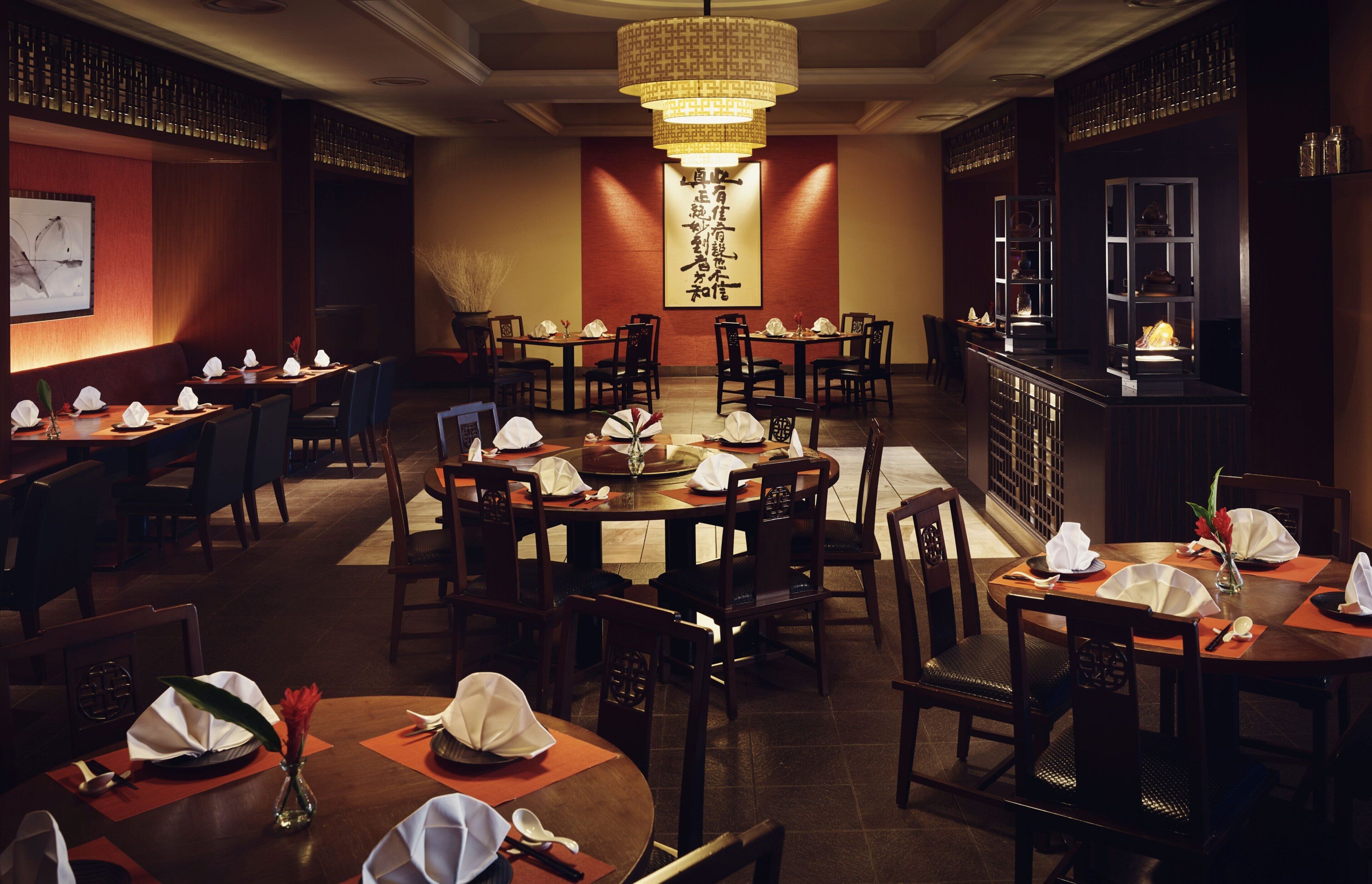 Un restaurante tradicional con varias mesas y sillas, una pared decorativa central con caligrafía, y una zona de bar con estanterías.