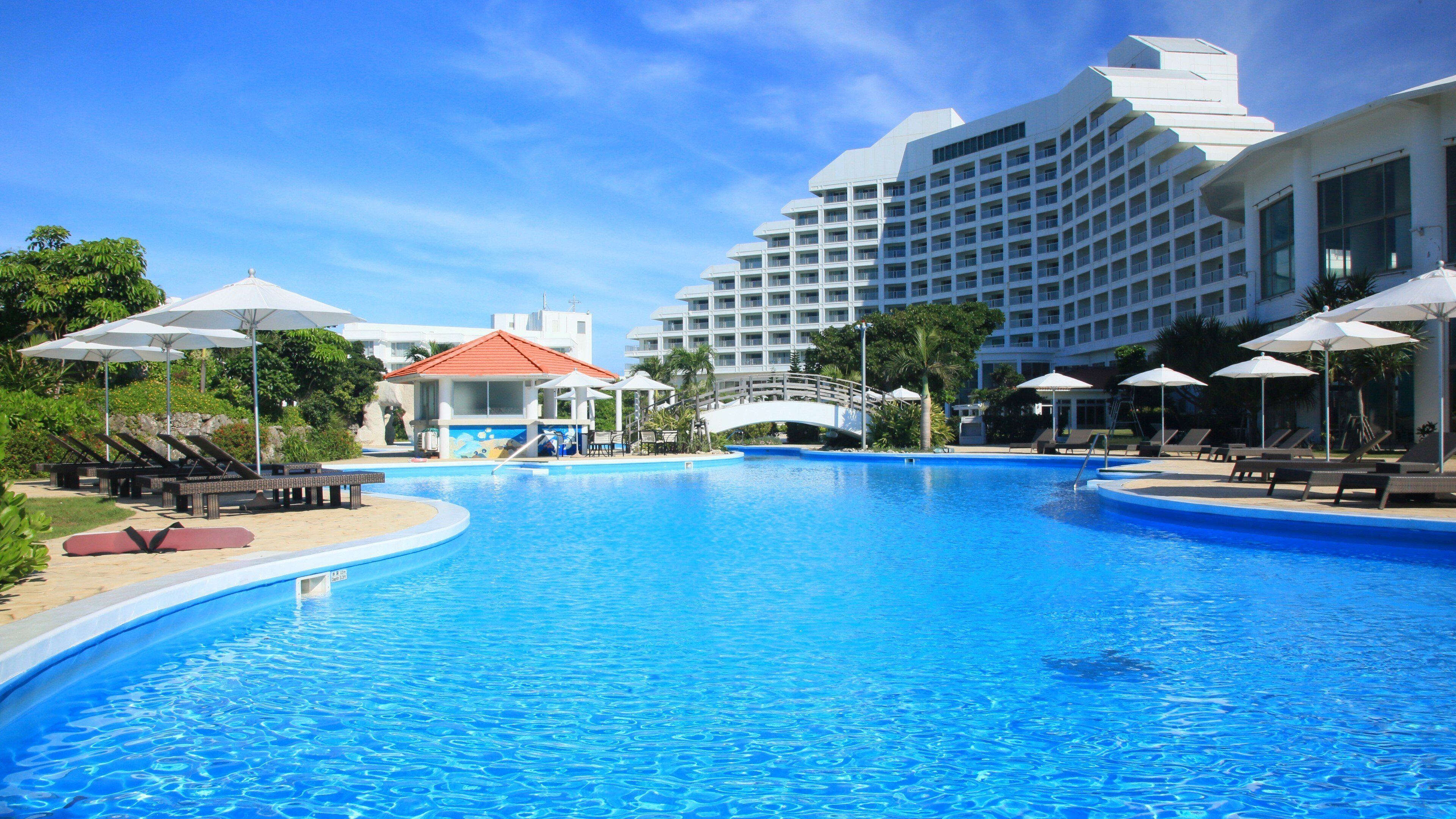 A large swimming pool with lounge chairs and umbrellas, surrounded by a hotel building with multiple floors and balconies.