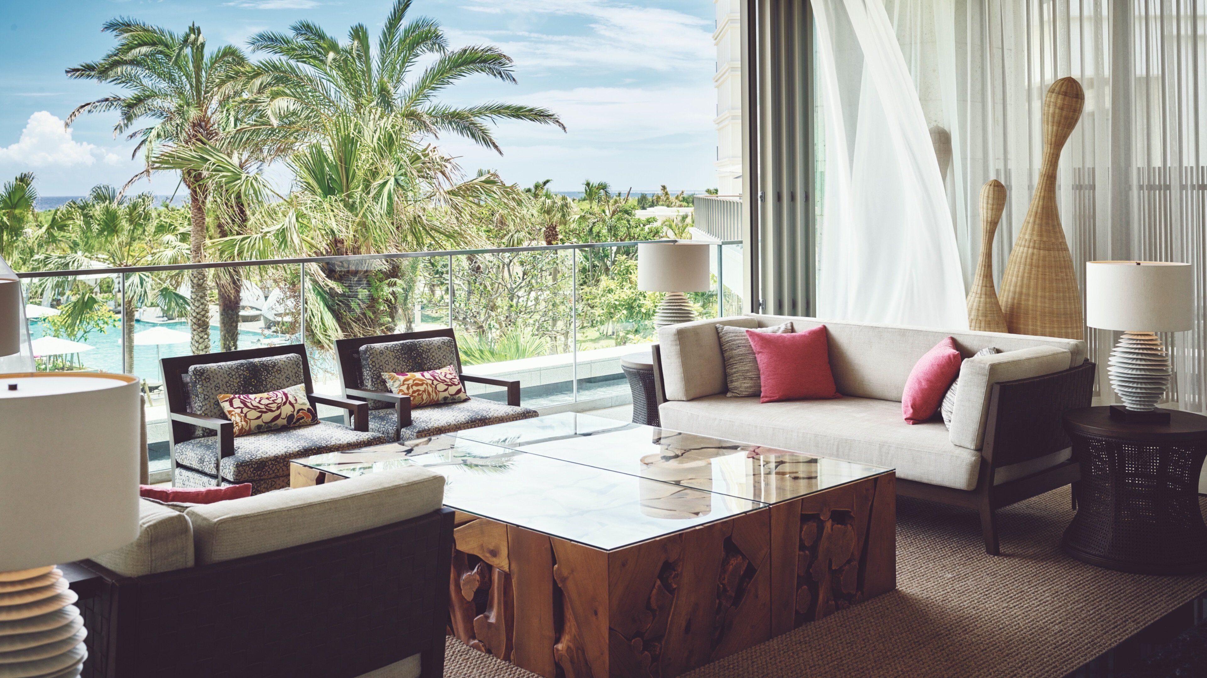 A modern living room with a glass coffee table, a sofa with cushions, and a view of a pool and palm trees.
