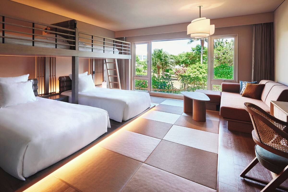 Une chambre d’hôtel moderne avec deux lits, un canapé, une petite table et un balcon donnant sur un espace verdoyant.