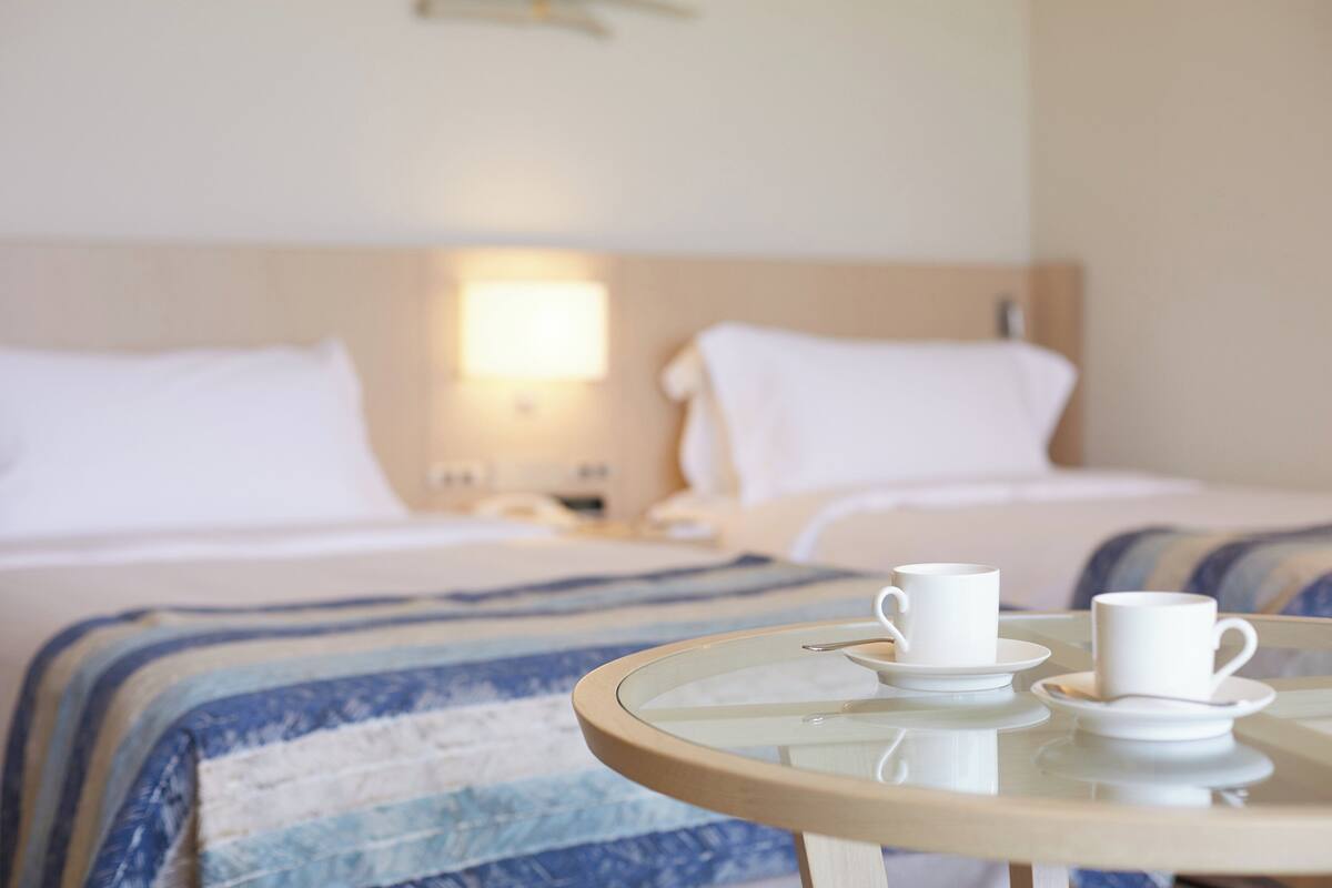 Une chambre d’hôtel avec deux lits, une table de chevet avec une lampe et une table ronde avec deux tasses et leurs soucoupes.