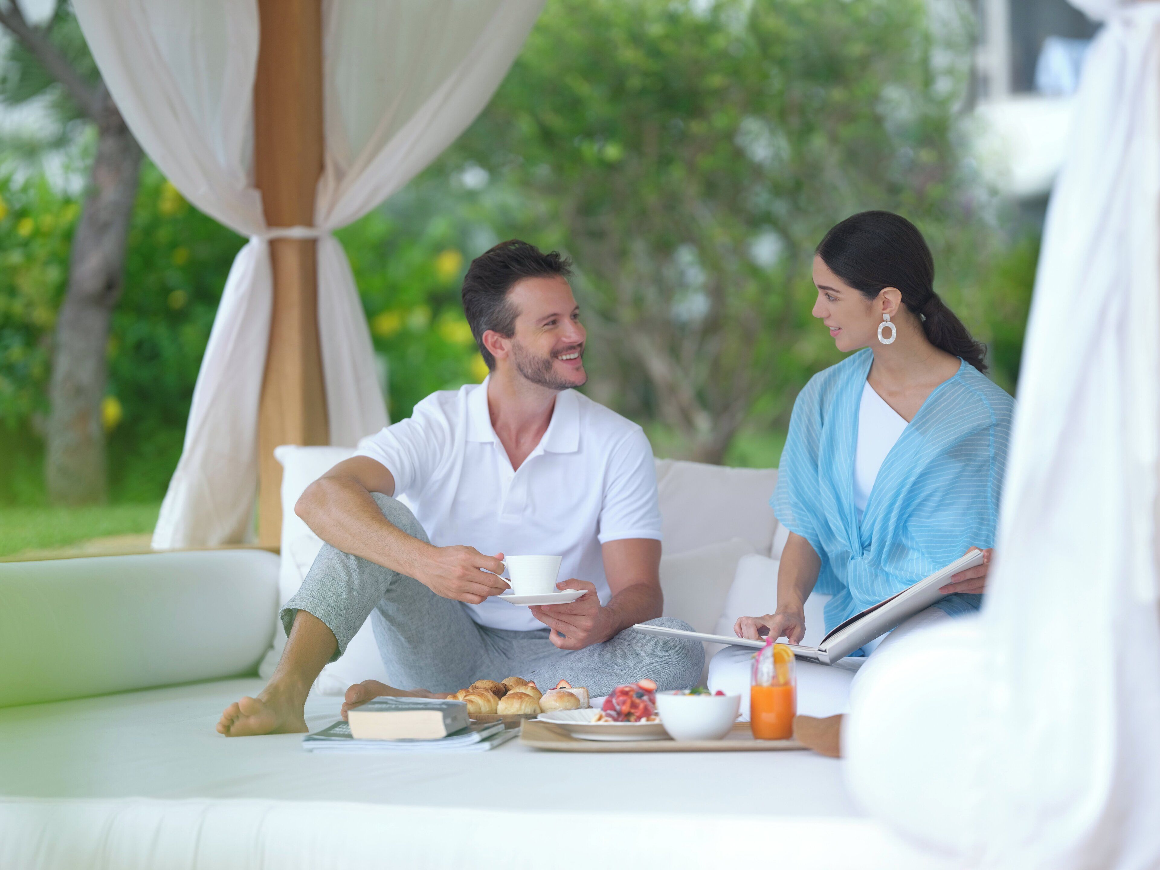 A couple sitting on a white sofa with a breakfast tray in front of them, including croissants, a bowl of fruit, and a glass of orange juice.