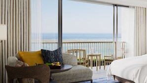 Une chambre d’hôtel avec un balcon donnant sur l’océan, un lit, un canapé avec des coussins, une petite table avec une lampe et une chaise.