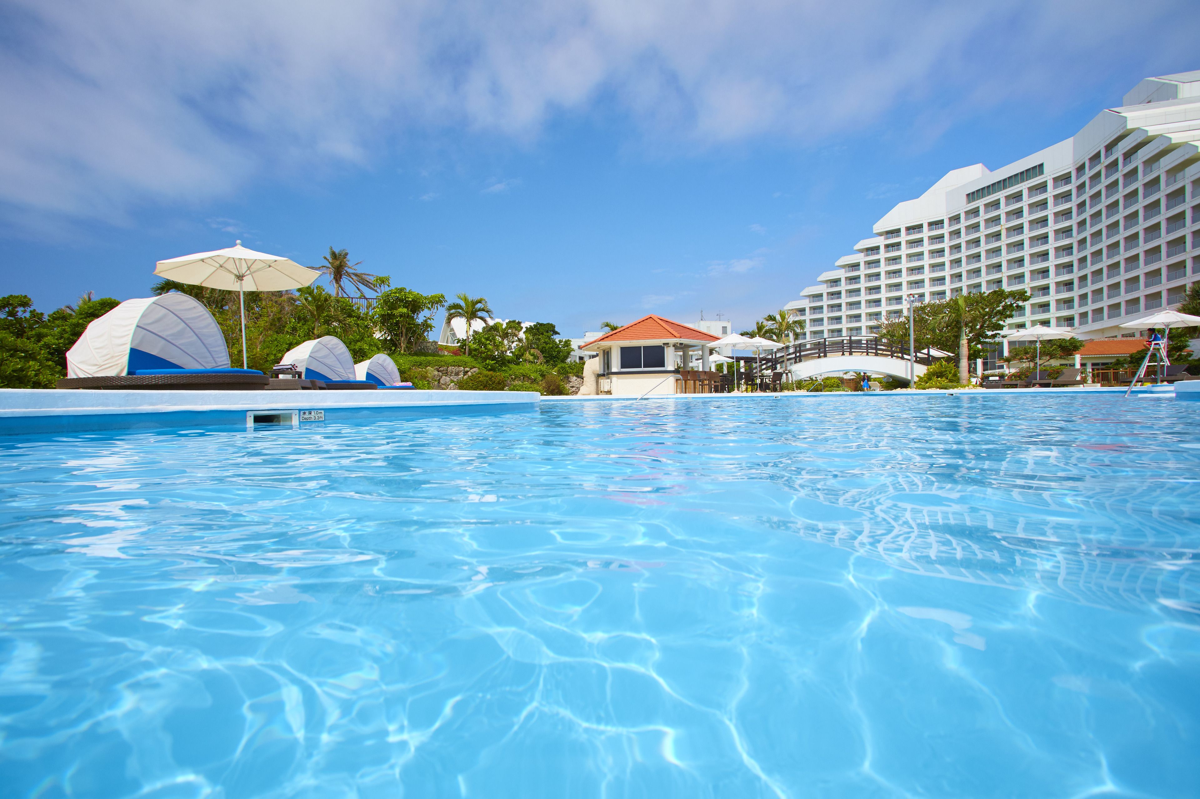 A swimming pool with clear blue water, surrounded by beach umbrellas and a large resort building.
