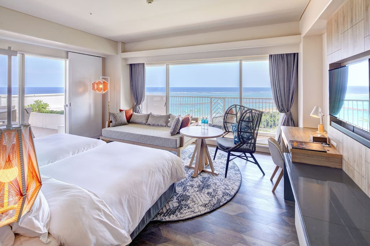 Une chambre d’hôtel avec deux lits, un balcon donnant sur la mer, une table ronde avec des chaises et un téléviseur.