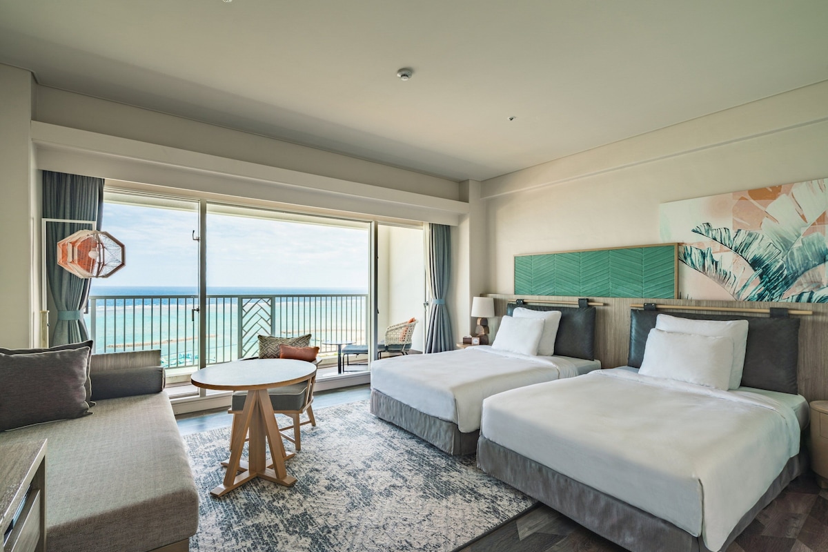 Habitación de hotel con dos camas, un sofá, una mesa de comedor y vista al mar.