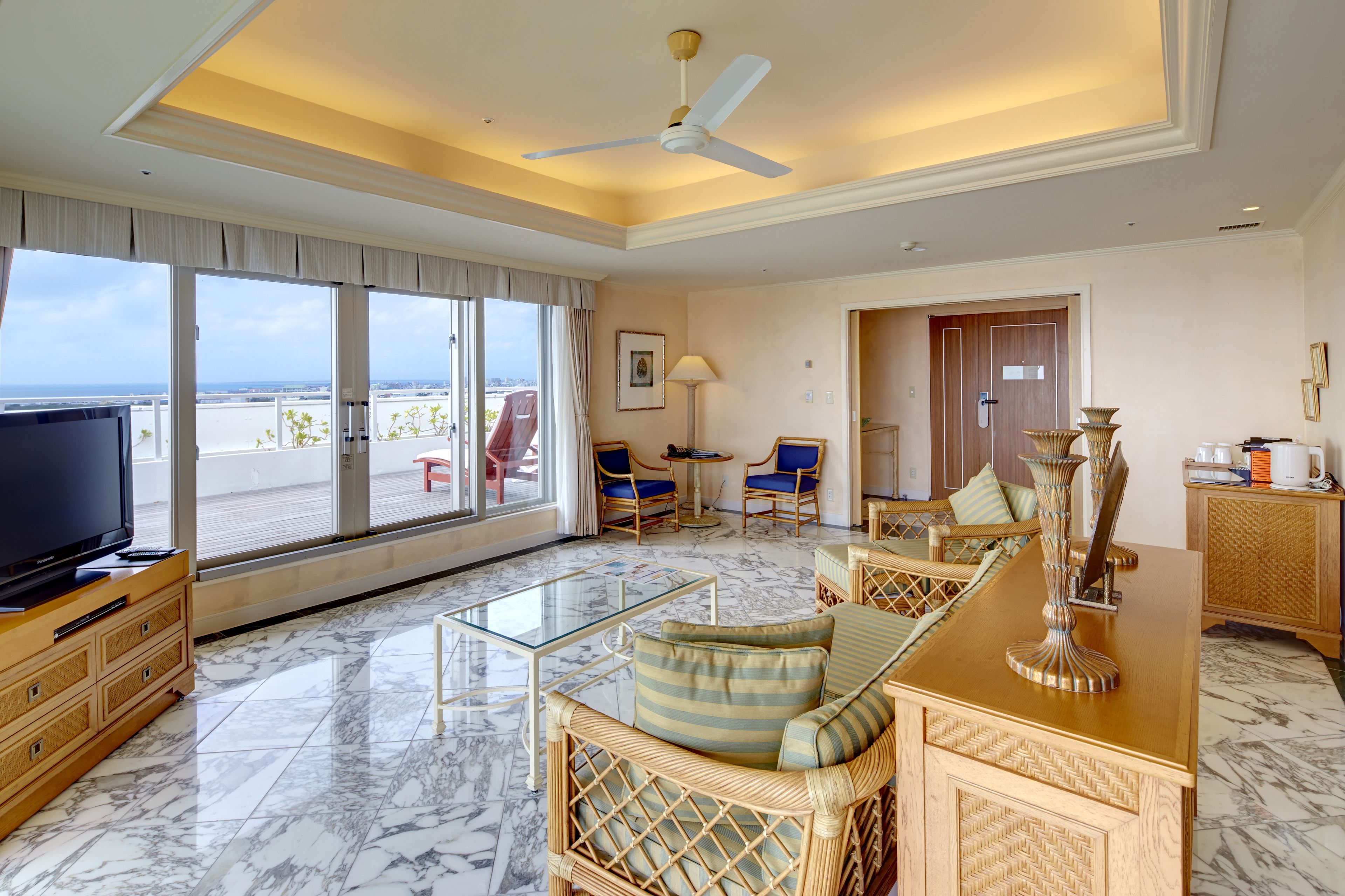A spacious living room with a large TV, comfortable seating, a glass coffee table, and a view of the beach through large windows.