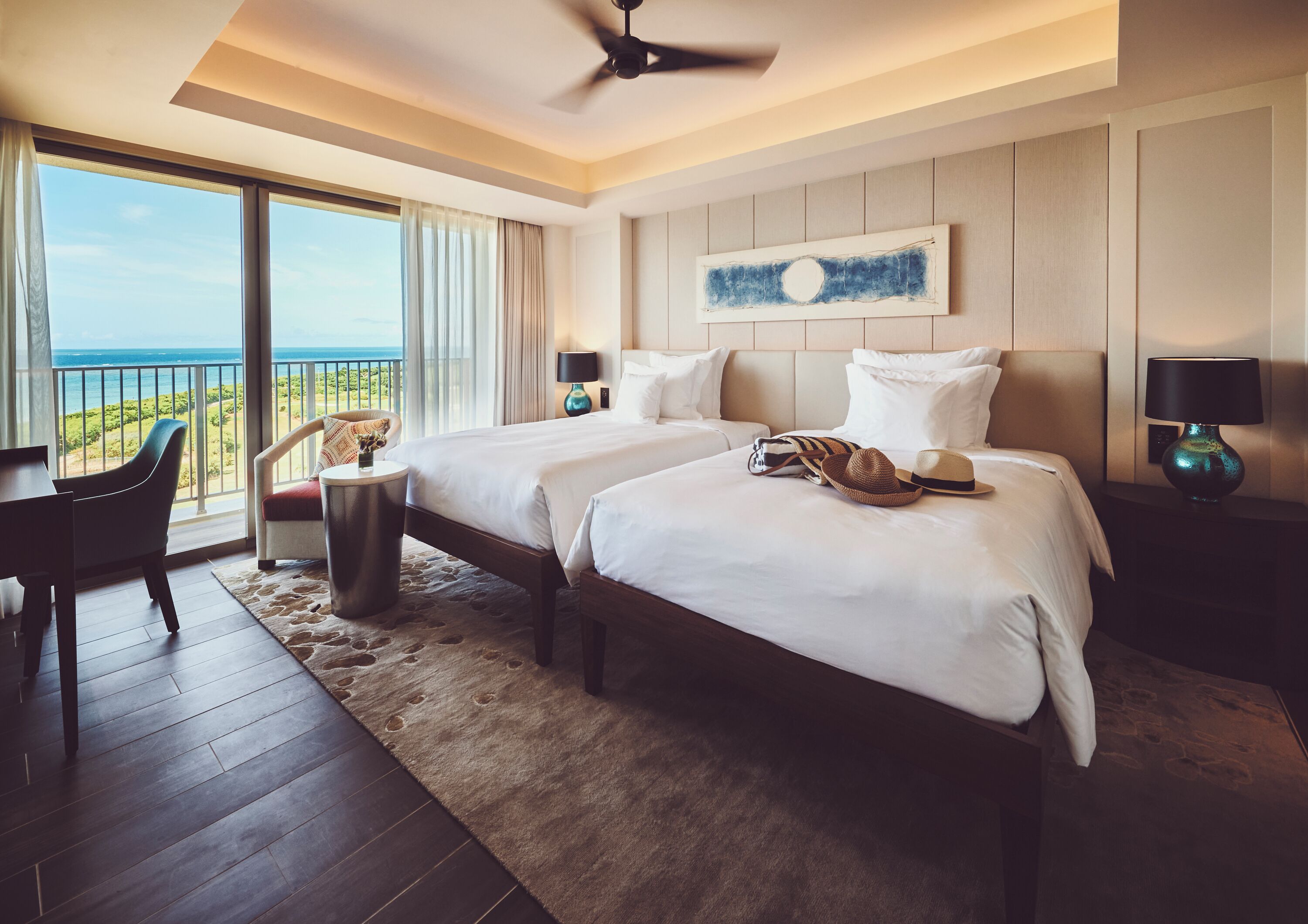 Ein Hotelzimmer mit zwei Betten, einem Schreibtisch und einem Sessel. Durch große Fenster ist der Blick aufs Meer.