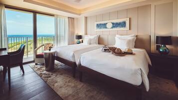 Ein Hotelzimmer mit zwei Betten, einem Schreibtisch und einem Sessel. Durch große Fenster ist der Blick aufs Meer.