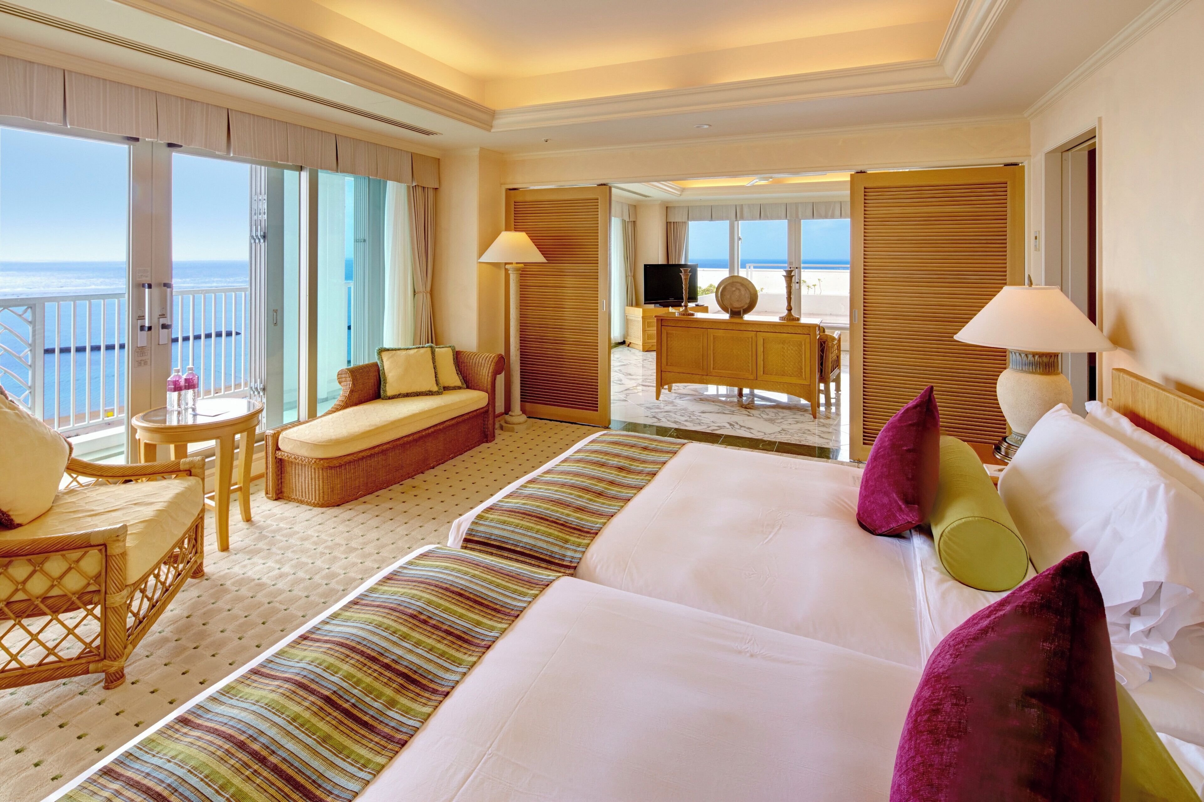 A hotel room with a large bed, a sofa, a desk with a TV, and a balcony with a view of the sea.