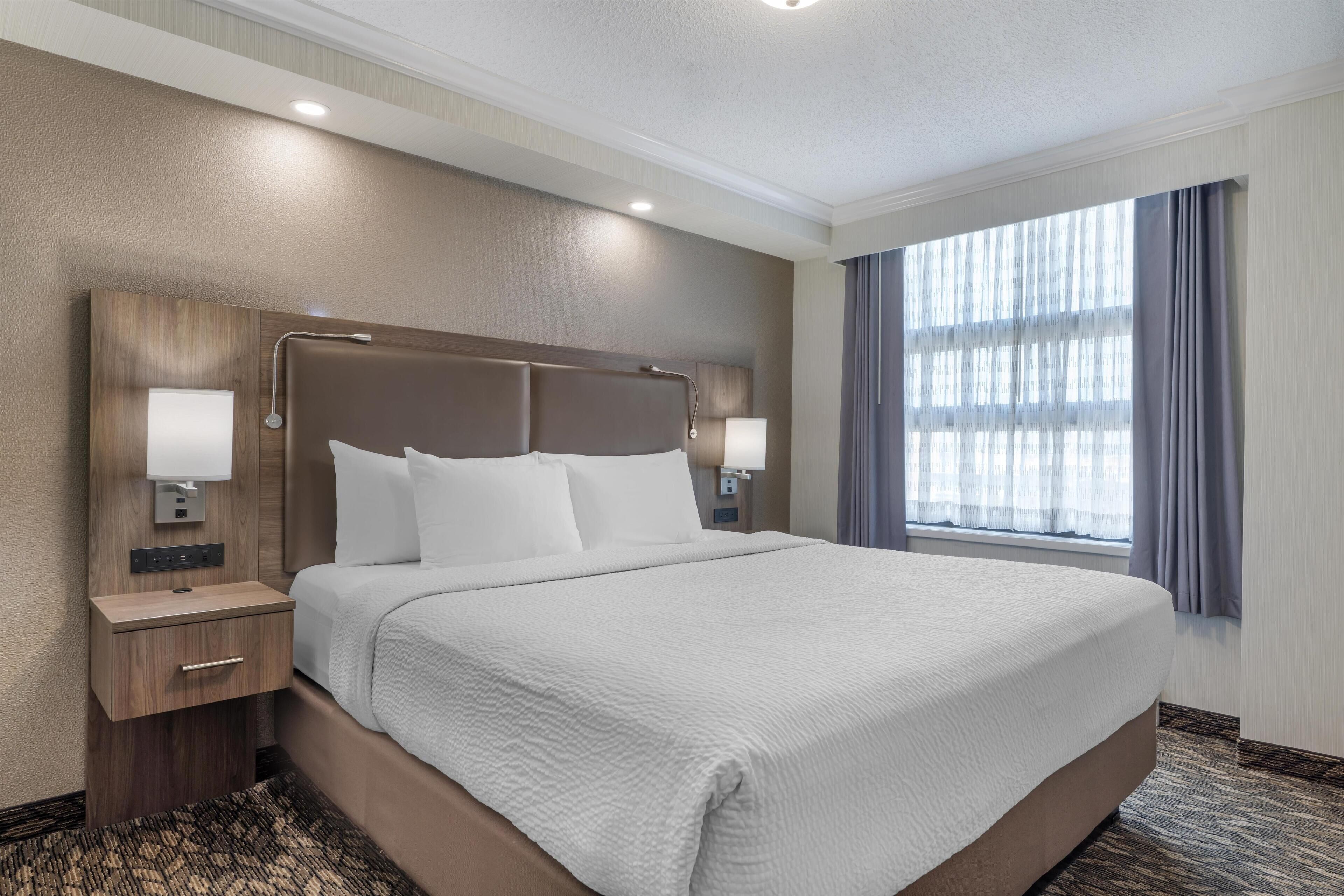 A hotel room with a large bed, two bedside tables with lamps, a wooden headboard, and a large window with curtains.
