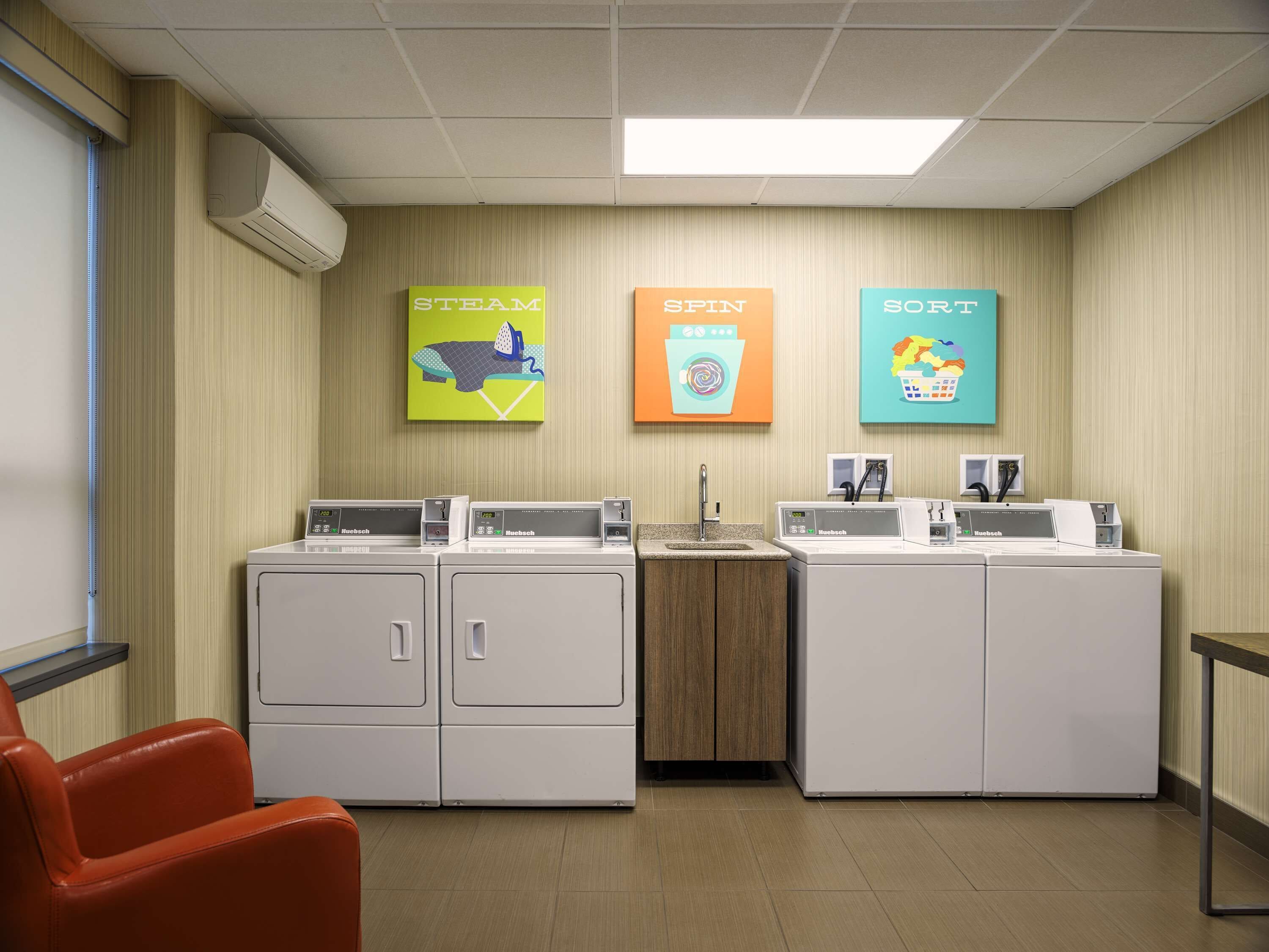 A laundry room with two rows of white front-loading washers and dryers, a wooden cabinet, and three colorful wall art pieces labeled "STEAM," "SPIN," and "SORT."